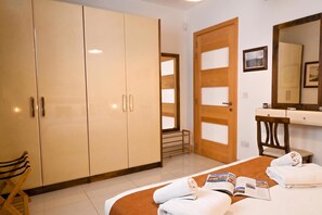 A hotel room with a bed, a bedside table with a magazine, a chair, a wardrobe, and a bathroom with a mirror and a towel rack. - Modern & Terrace by the Beach, 3brd in Qawra (St. Paul's Bay)