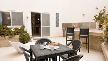 Un patio con mesa y sillas dispuestas para dos, una pequeña cocina y un ventanal corredero que da a un espacio interior.