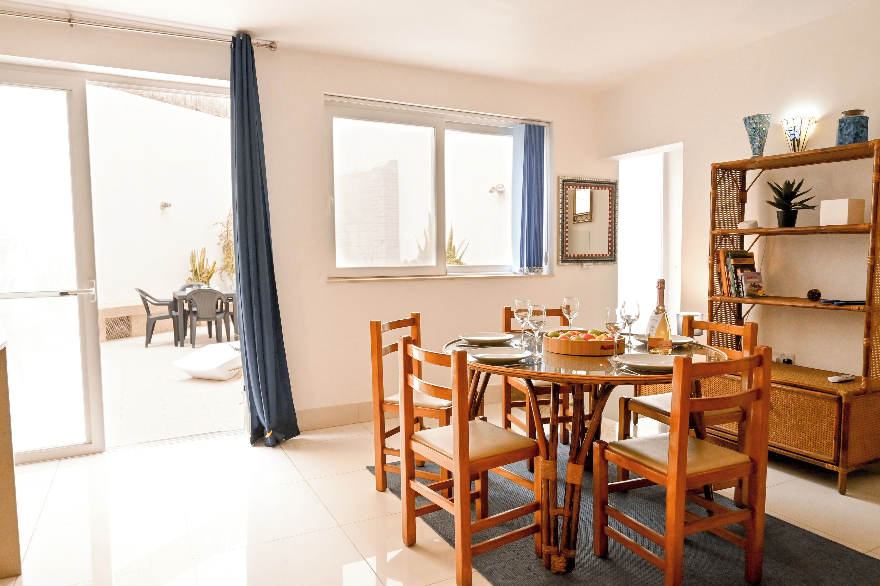 Un coin repas avec une table dressée pour quatre personnes, des chaises en bois, une bibliothèque et une grande fenêtre avec des rideaux.