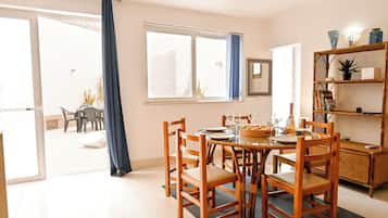 Un comedor con una mesa dispuesta para cuatro, sillas de madera, una estantería y un gran ventanal con cortinas.