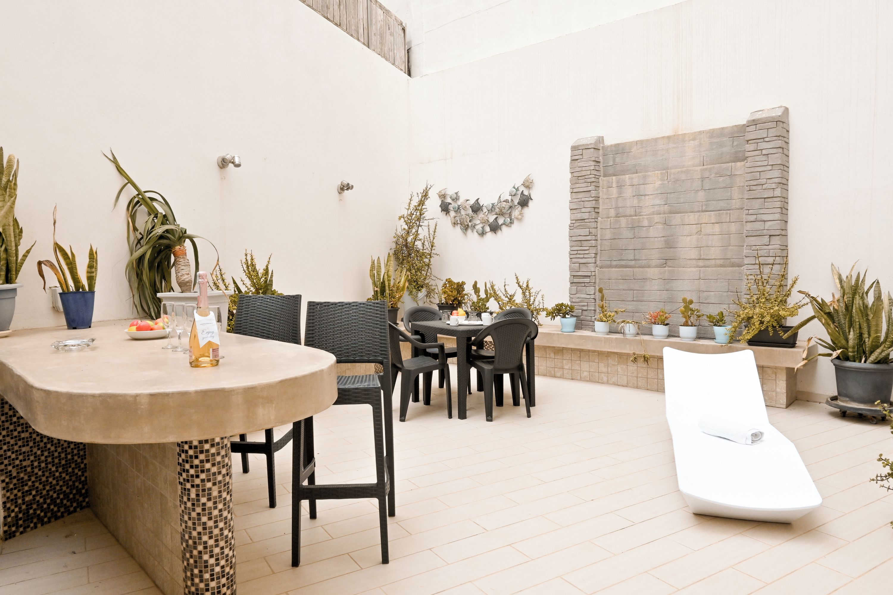 Un patio al aire libre con una mesa redonda, sillas, una tumbona y varias plantas en macetas.