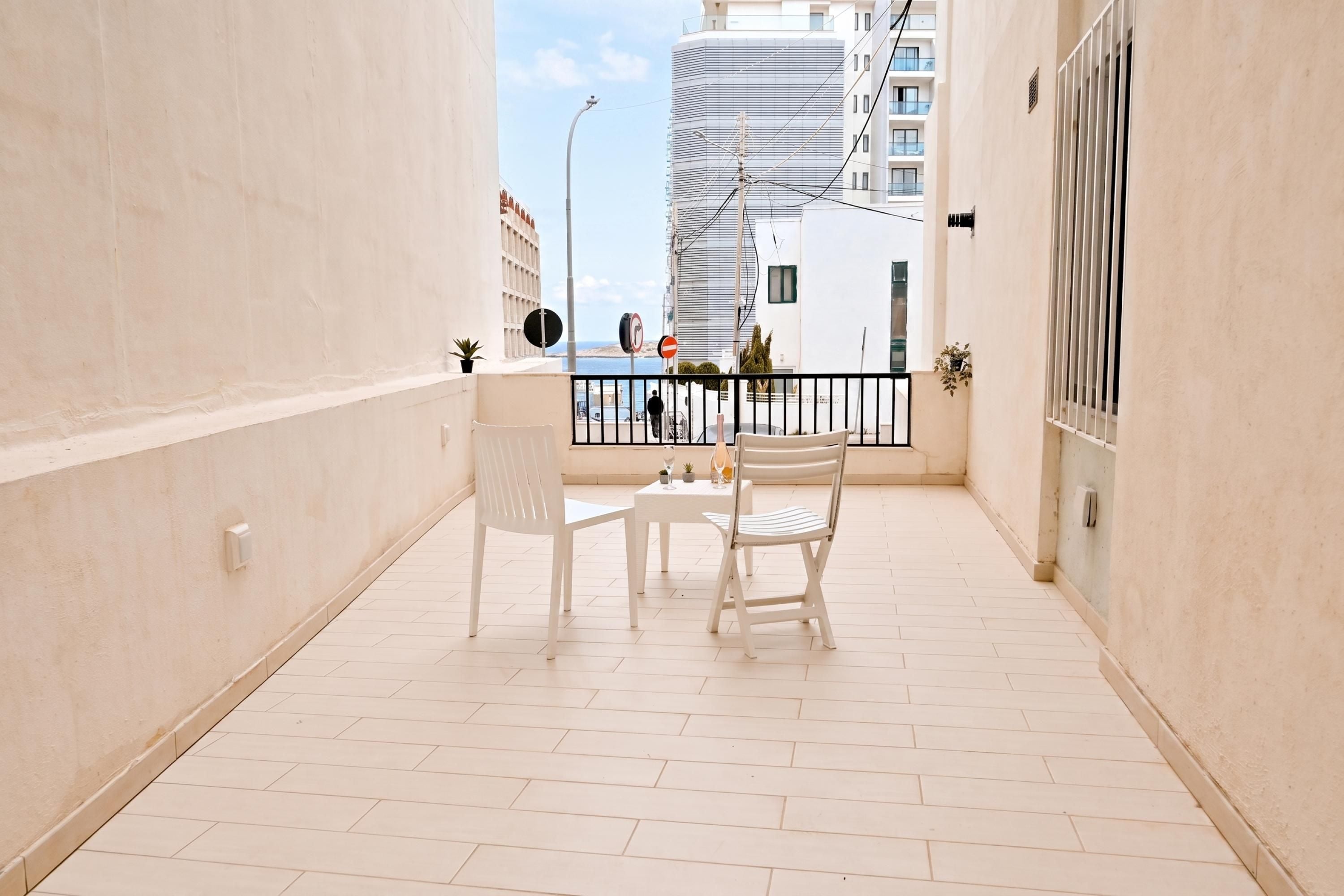 A balcony with a white table and chairs, a view of the sea, and a building in the background.