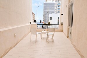 Un balcón con una mesa y sillas blancas, vista al mar y un edificio en el fondo.