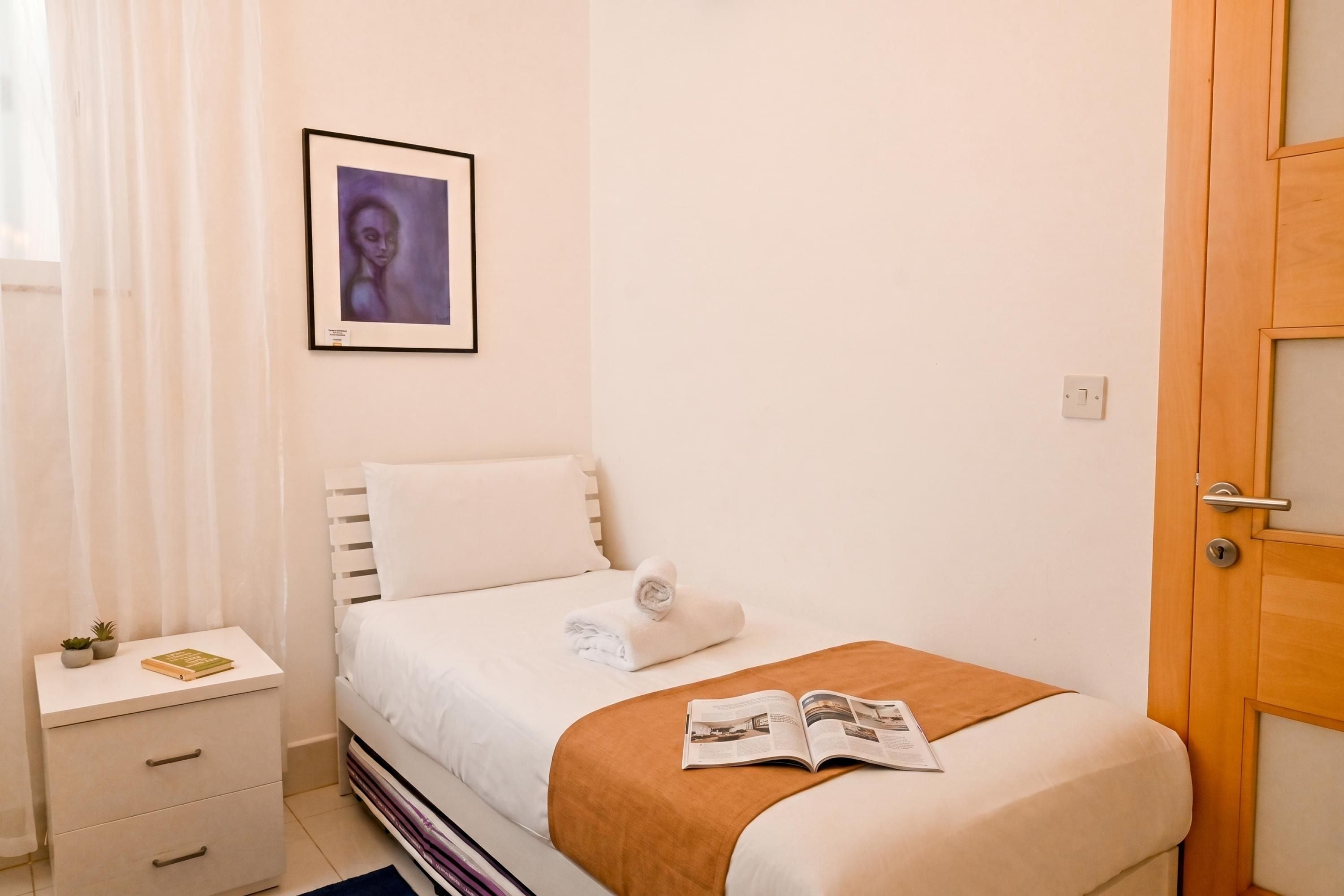 Una cama bien tendida con una colcha blanca y una manta marrón, una mesita de noche con un libro y una planta pequeña, y un cuadro en la pared.