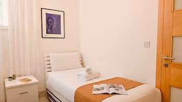 Una cama bien tendida con una colcha blanca y una manta marrón, una mesita de noche con un libro y una planta pequeña, y un cuadro en la pared.