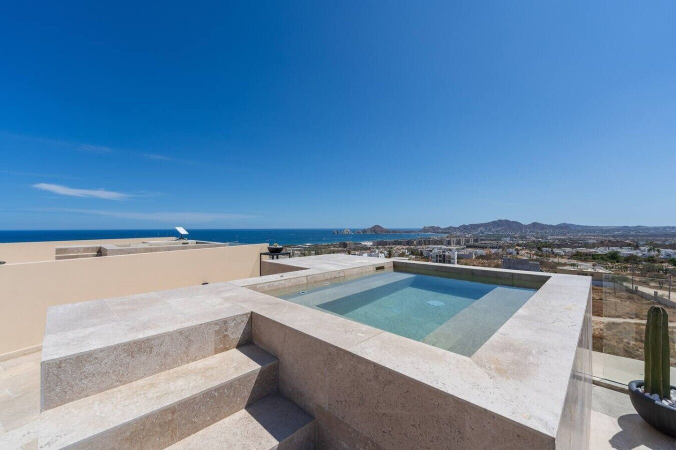 Une piscine sur le toit, avec une eau d’un bleu cristallin, entourée d’escaliers et offrant une vue sur l’océan et les collines lointaines.