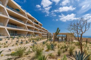 Um hotel moderno com várias varandas, um jardim desértico com cactos e vista para o oceano.