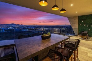 A modern dining area with a panoramic city view at dusk, featuring a long table and chairs, a potted plant, and pendant lighting. - Luxury penthouse with stunning ocean and Arch views. Features a rooftop deck with private jetted plunge pool, grill, ping pong table, and Starlink WiFi. Near beaches, top restaurants, shopping, and Cabo nightlife for the perfect getaway. (Cabo San Lucas)