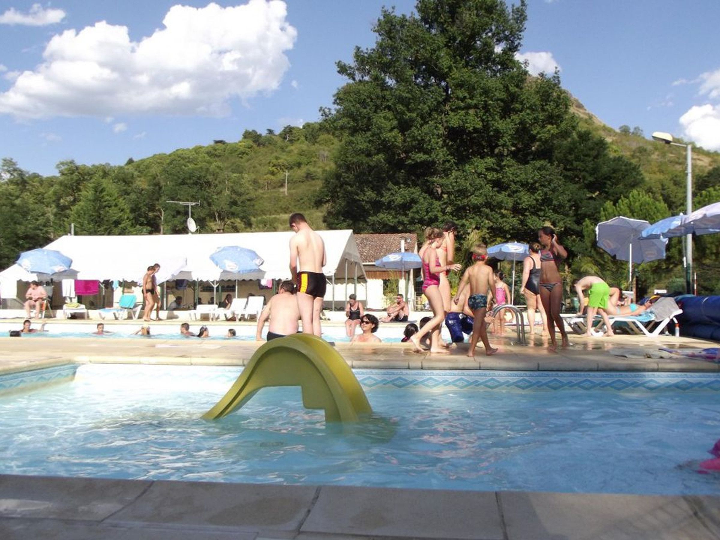 A swimming pool with people around it, a hill with trees in the background, and a building with a thatched roof.