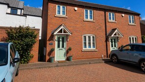 A two-story brick house with a green door and white window frames, a driveway, and two cars parked in front. - Central Bridgnorth Boutique Townhouse - Private Parking (Bridgnorth)