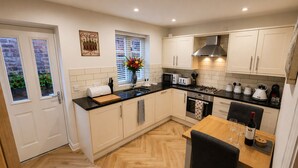 A compact kitchen with white cabinets, a black countertop, a stove, a microwave, and a dining area with a table and chairs. - Central Bridgnorth Boutique Townhouse - Private Parking (Bridgnorth)