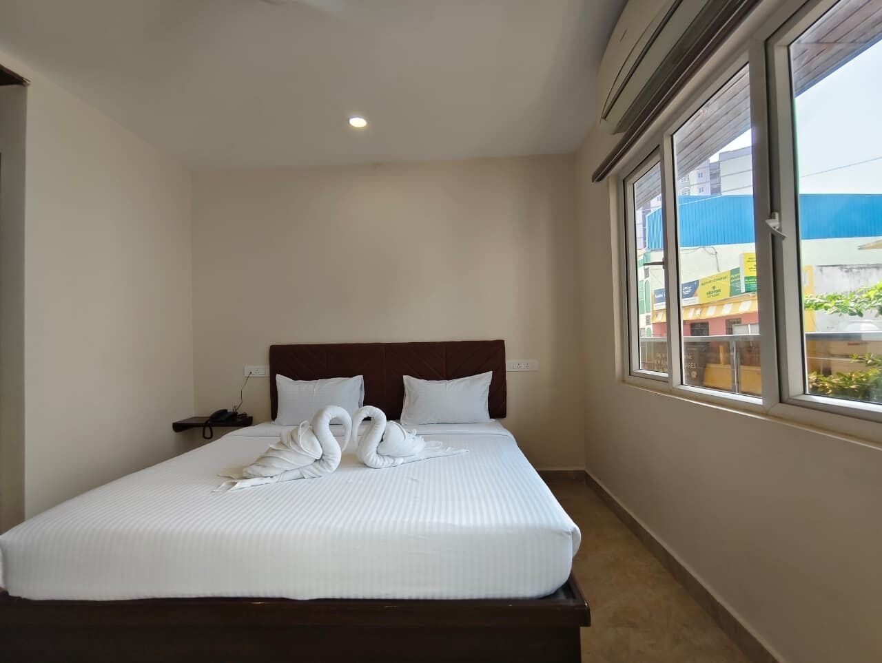 A hotel room with a large bed, a wooden headboard, a bedside table with a phone, and a window with a view of buildings.