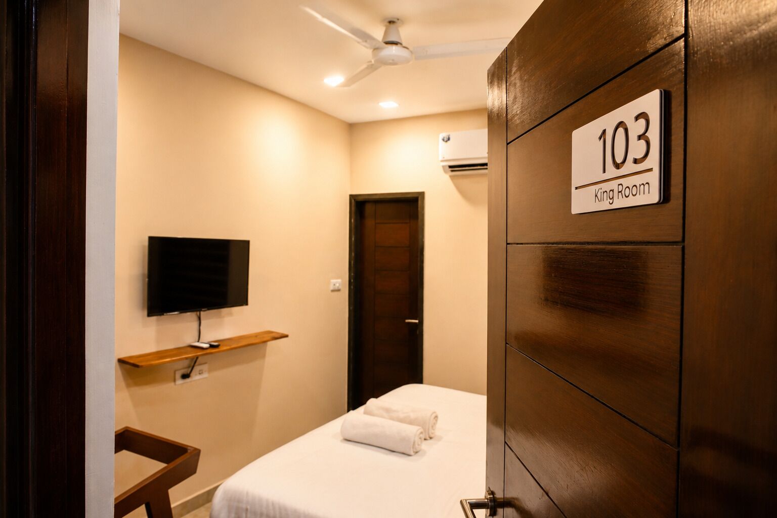 A hotel room with a bed, a TV mounted on the wall, a wooden door, and a sign indicating room number 103.