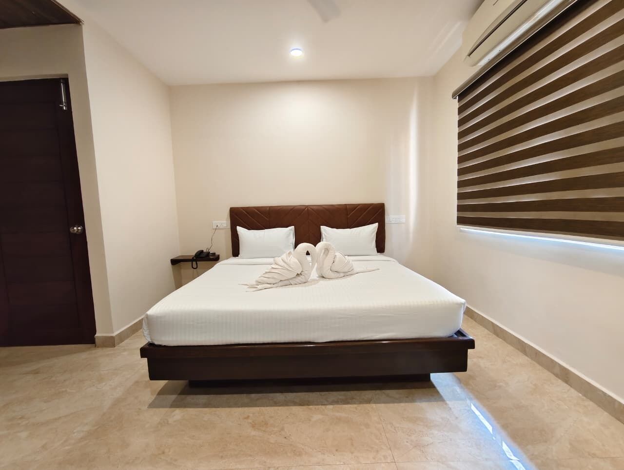 A neatly made bed with a wooden headboard, a bedside table with a lamp, and a window with blinds.