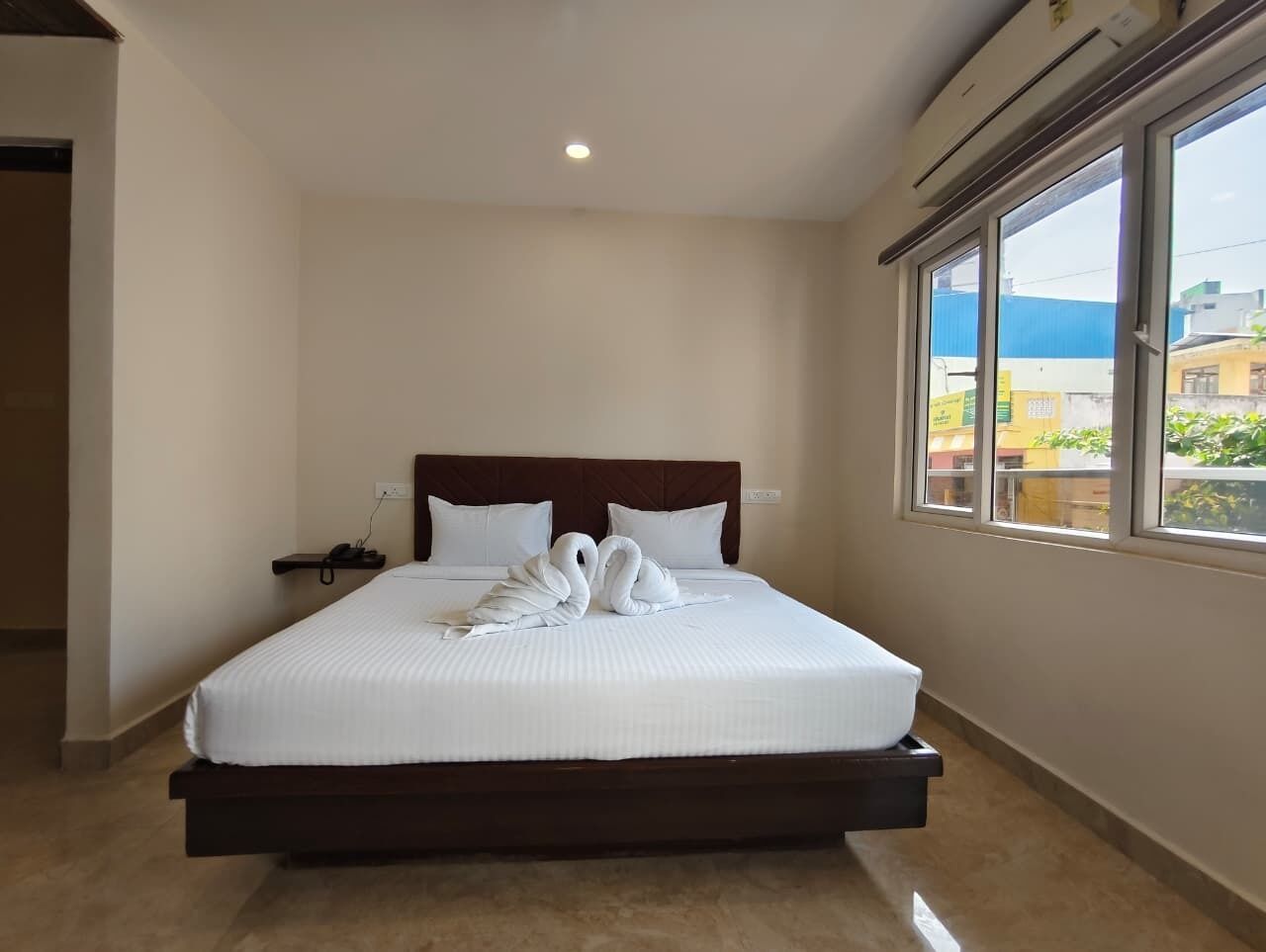 A hotel room with a large bed, a headboard, a bedside table, and a window with a view of buildings.