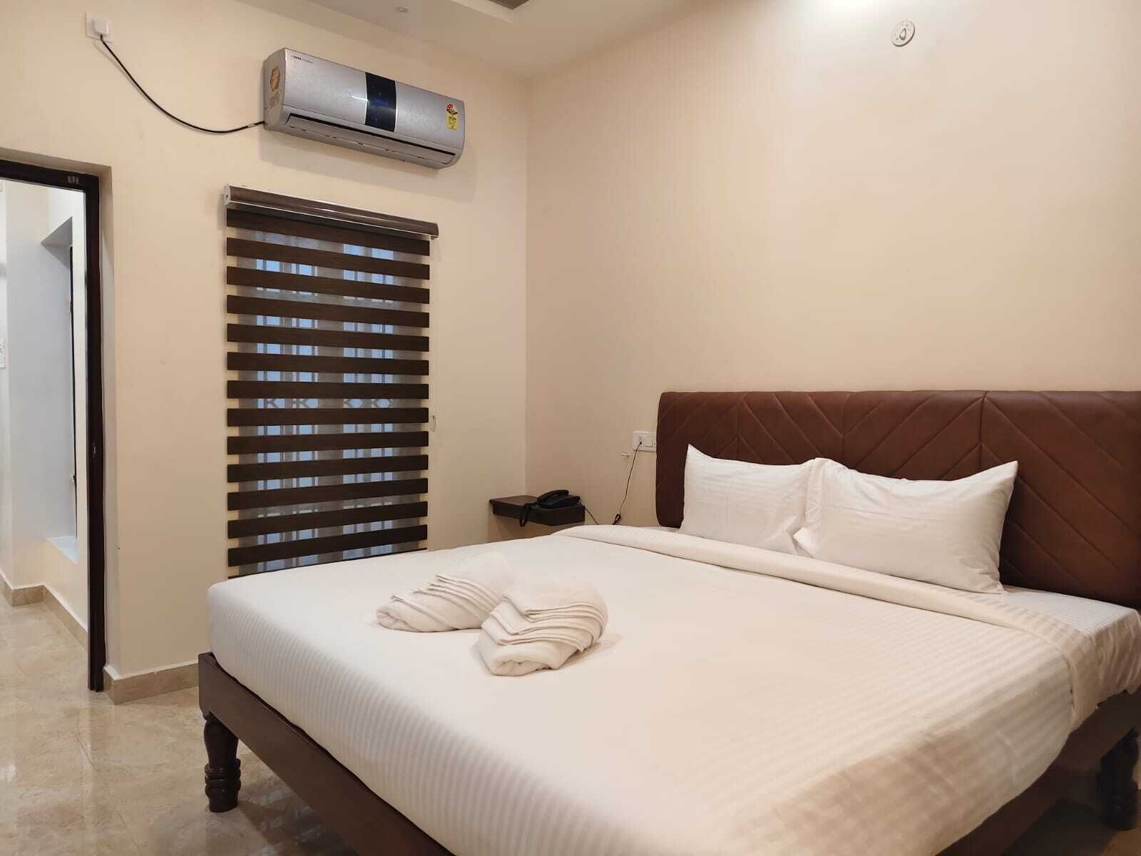 A hotel room with a bed, a wall-mounted air conditioner, a window with blinds, and a bedside table with a phone.