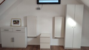 Quarto com teto inclinado, uma cama, guarda-roupa, penteadeira, radiador e televisão montada na parede.