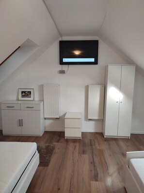 A bedroom with a sloped ceiling, a bed, a wardrobe, a dresser, a radiator, and a television mounted on the wall.
