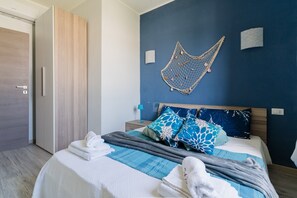 A neatly made bed with blue and white bedding, a wooden wardrobe, and a hammock on the wall. - Mediterranea House A Otranto – 8 Guests, Balcony, Wi-Fi & Air Conditioning (Otranto)