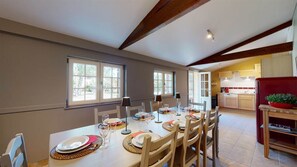 Un coin repas avec une table dressée pour six personnes, une cuisine en arrière-plan et de grandes fenêtres.