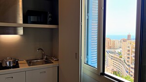 A kitchen with a sink, stove, and cabinets, and a window with a city view. - Appartement 6A - MyResidenced'Azur (Beausoleil)