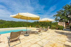 A poolside area with lounge chairs and umbrellas, a pool, and a view of a forested hill.