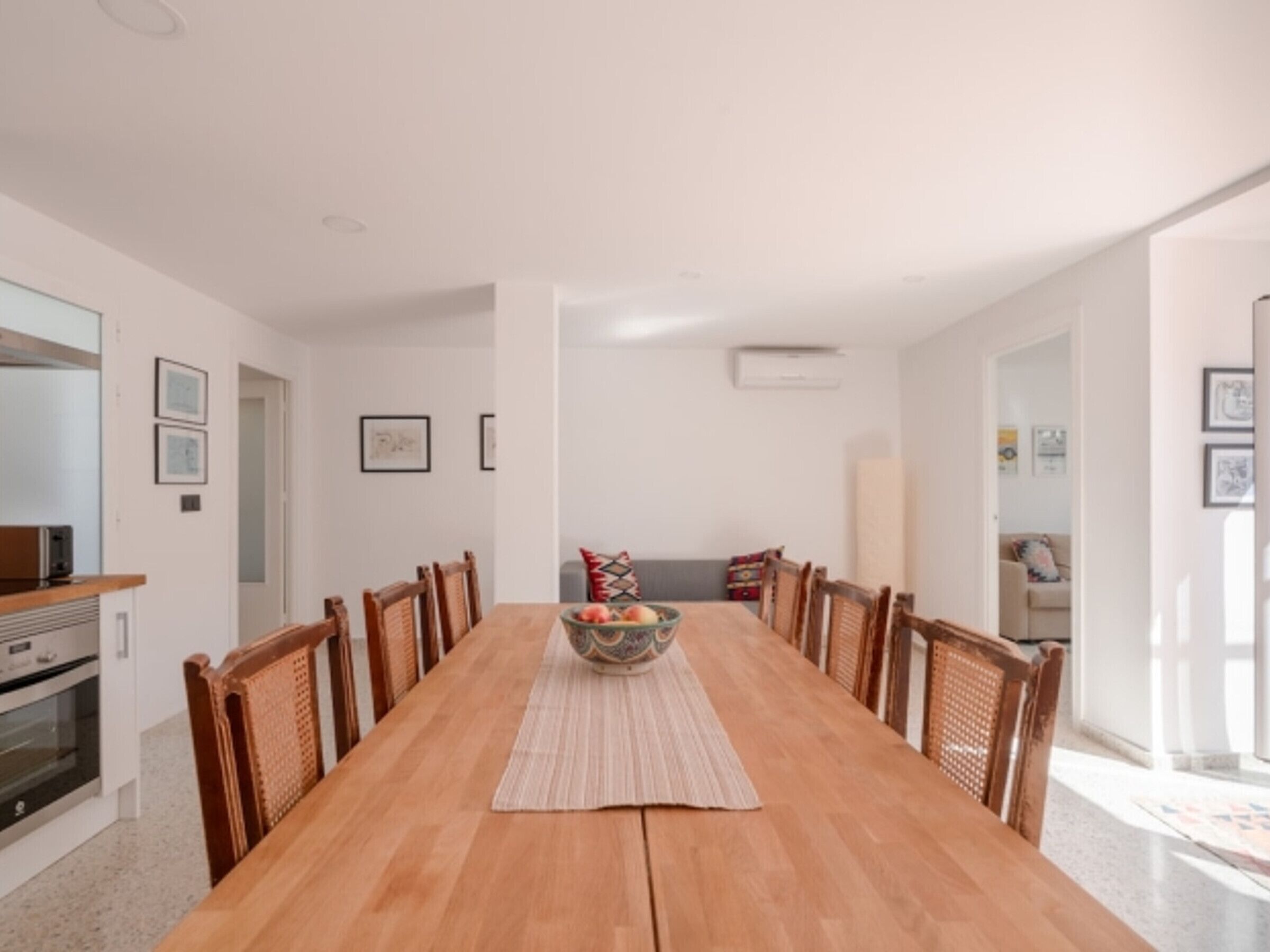 A long wooden dining table with chairs, a central woven placemat, and a bowl of fruit.