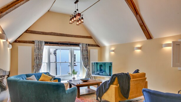 Ein Loft-Zimmer mit schrägem Dach, großem Fenster mit Vorhängen, Fernseher und gemütlicher Sitzecke mit Sofa und Sesseln.