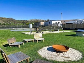 Ein Garten mit Feuerstelle, Holzbänken, Schaukel und umzäunter Fläche.