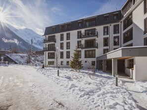 Ein modernes, mehrstöckiges Gebäude mit Balkonen, Schnee auf dem Boden und Bergen im Hintergrund.