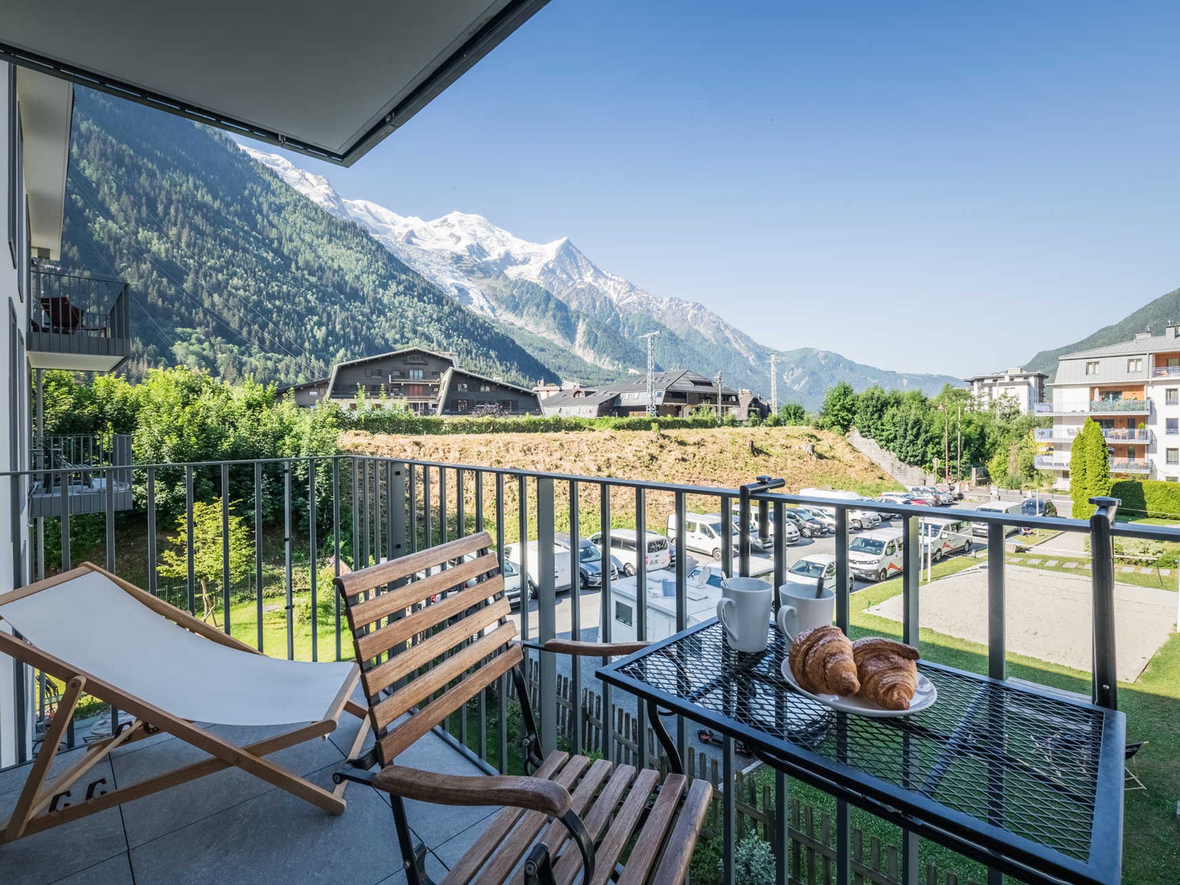 A balcony with a wooden bench, a metal table with croissants, and a view of snow-capped mountains.