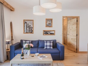 Ein Wohnzimmer mit einem blauen Sofa, einem Couchtisch aus Holz mit einer Flasche und Gläsern sowie gerahmten Bildern an der Wand.