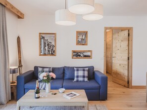 Een woonkamer met een blauwe bank, een bijzettafeltje met een fles en glazen, en twee ingelijste foto's aan de muur.