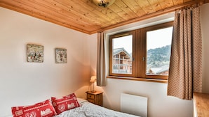 A bedroom with a wooden ceiling, a bed with white bedding and red pillows, a nightstand with a lamp, and a window with curtains. - Spacious apartment near slopes - 6 guests (Les Gets)