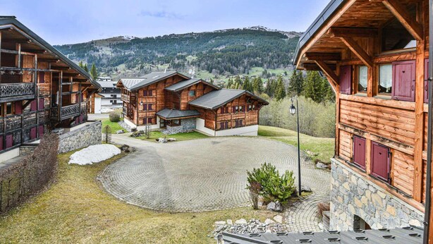A wooden chalet-style building with a stone foundation, a paved courtyard, and a view of a mountainous landscape. - Spacious apartment near slopes - 6 guests (Les Gets)
