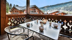A wooden deck with a table set for dining, overlooking a mountainous landscape with traditional wooden buildings. - Spacious apartment near slopes - 6 guests (Les Gets)
