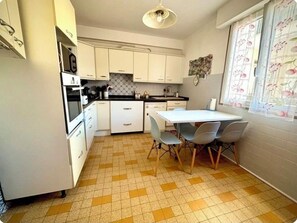 A compact kitchen with white cabinets, a tiled floor, a dining table with chairs, and a window with a floral curtain. - At the gates of Cannes: Apartment 100m2 and terrace, center, Croisette on foot (Le Cannet)