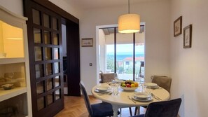 Ein Essbereich mit einem runden Tisch, gedeckt für vier Personen, einem großen Fenster mit Meerblick und einer dunklen Holztür mit Glasfeldern.