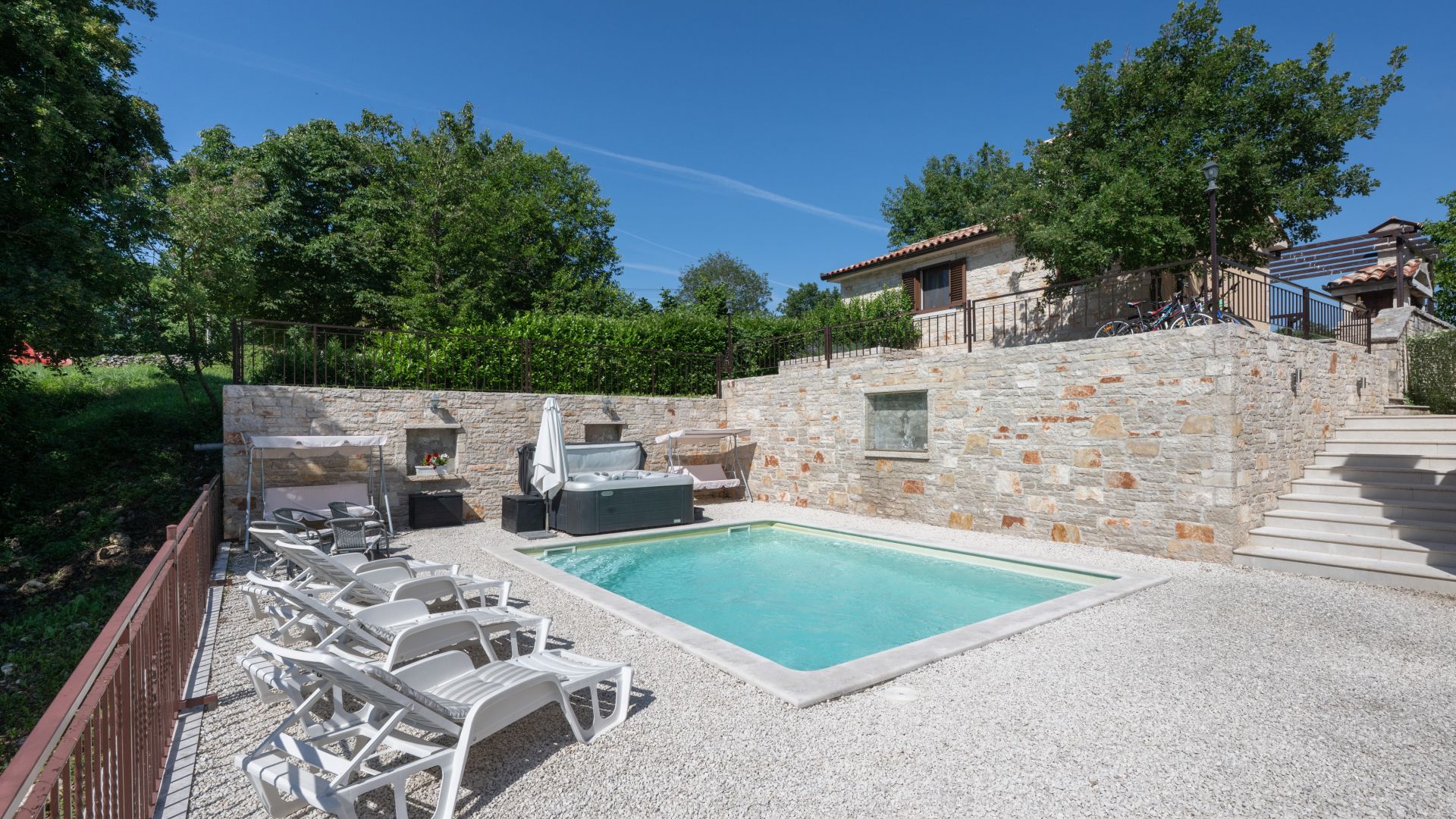 A swimming pool with lounge chairs, a hot tub, and a stone wall.