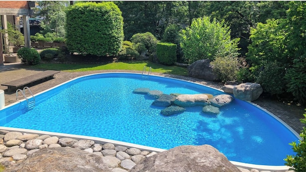 A swimming pool with a stone border and rocks inside, surrounded by greenery and a brick structure.