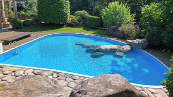 En swimmingpool med en stenkant og sten indeni, omgivet af grønt og en murstensbygning.