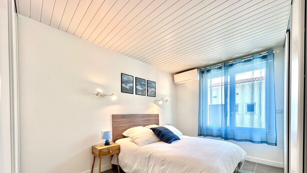Un dormitorio con cama, mesita de noche, luces fijadas a la pared, ventana con cortinas y suelo de baldosas.
