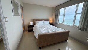 A bedroom with a bed, nightstands, a lamp, a view of the sea, and a window with curtains. - Oceanfront two-bedroom (Surfside)
