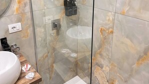 A bathroom with marble tiles, a glass shower enclosure, a white sink, and toiletries on a wooden shelf. - Donna Flora's House (Napoli)