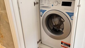 A compact washing machine in a small room with a wooden shelf above it. - Donna Flora's House (Napoli)