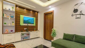 A modern living room with a flat-screen TV, wooden panel wall, white shelving with decorative items, a green sofa, and a black coffee table. - Tucson 2BHK Forest view by Homeyhuts (Dehradun)