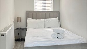 Une chambre à coucher avec un lit, une fenêtre avec des stores, une table de chevet avec une lampe et un radiateur.