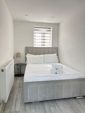 Ein Schlafzimmer mit einem Bett, einem Fenster mit Jalousien, einem Nachttisch mit Lampe und einem Heizkörper.