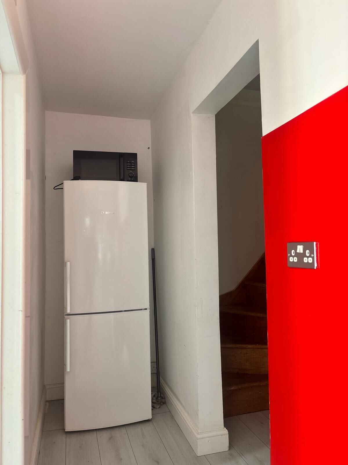 A compact kitchen with a white refrigerator and a microwave, a broom leaning against the wall, and a red door.