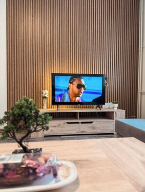 Ein Fernseher auf einem Holzständer in einem Raum mit einer Lamellenwand, einer kleinen Pflanze und einem dekorativen Gegenstand auf dem Ständer.